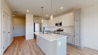 This well-appointed single-floor residence offers a thoughtfully designed layout across seven rooms, blending modern finishes with functional living spaces throughout. The kitchen serves as a central highlight, featuring a large central island with a white quartz countertop, an undermount sink with a matte black faucet, and stylish pendant lighting overhead. Shaker-style cabinetry in crisp white and soft gray tones lines the walls, complemented by a classic white subway tile backsplash and a full suite of stainless steel appliances, including a gas range, microwave, and refrigerator. Light hardwood-style flooring flows through the main living and kitchen areas, creating a warm, cohesive aesthetic. The bedrooms feature plush carpeting and large windows that invite abundant natural light and offer views of the surrounding building exterior. One bedroom is further enhanced by a ceiling fan. The bathroom includes a full soaking tub and shower combination with brushed nickel fixtures set against clean white surrounds. A dedicated in-unit laundry closet houses a full-size washer and dryer side by side. Throughout, the interior maintains a bright, neutral palette with white walls and clean architectural lines, contributing to an open and modern ambiance.