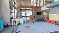 This contemporary community clubhouse is designed as a dynamic, multi-functional social space spread across a single open floor plan. The interior is characterized by soaring ceilings, large floor-to-ceiling windows, and an abundance of natural light that floods in from multiple directions, offering views of the surrounding courtyard and outdoor amenity areas.

The space is defined by its bold mid-century modern aesthetic, featuring a vibrant palette of teal, orange, red, and yellow accents contrasted against neutral grays and warm wood ceiling panels. A striking sputnik-style chandelier anchors the main lounge area, complemented by pendant lighting throughout.

A dedicated game area houses a full-size pool table beneath expansive windows, while a built-in bar with colorful stools provides a social gathering point. The lounge zone centers around a double-sided tile fireplace with a wall-mounted flat-screen television, flanked by plush sofas, accent chairs, and striped booth-style seating.

Additional areas include a reception and workstation zone with an illuminated mosaic accent wall, casual dining tables with modern seating, and intimate conversation nooks with tufted upholstered panels. Throughout, contemporary artwork and sculptural décor elements reinforce the playful, design-forward character of the space.