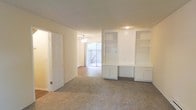 This two-story residence offers a clean, neutral interior designed for comfortable everyday living. The main living area is finished with plush carpeting and painted in soft, warm tones that create a bright and welcoming atmosphere throughout. A standout feature of the living space is the built-in wall unit, which includes open shelving above and enclosed cabinetry below — providing generous storage and display options while adding a sense of architectural character to the room.

The layout flows openly between the living and adjoining areas, with a ceiling fan-lit dining or transitional space visible beyond. A sliding glass door or large window at the rear of this adjoining room invites natural light deep into the interior and provides direct access to an outdoor patio or balcony area, offering views of the exterior surroundings.

Hardwood-style flooring transitions the space near the entry and adjoining rooms, complementing the carpeted living area. A baseboard heater is present along the wall, indicating a functional and efficient heating setup. The overall design is understated and move-in ready, with a neutral palette suited to a variety of furnishing styles.