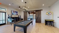 This single-story social lounge in Spring, TX, is thoughtfully designed to accommodate a wide range of recreational and social activities. The space is anchored by a spacious great room featuring dramatic vaulted ceilings with rich, dark exposed wood beams and tongue-and-groove wood detailing overhead. Light hardwood flooring runs throughout, lending warmth and visual continuity across the entire layout. A large chandelier illuminates a generous dining area, while full-height glass doors open to an outdoor view, filling the room with natural light. An adjacent bar and kitchen area includes a lengthy counter with seating, pendant lighting, and a built-in beverage station with mosaic tile backsplash and under-counter refrigeration.

The game room is separated from the great room by a striking pair of sliding barn doors in distressed wood. Inside, a full-size billiard table sits beneath an industrial-style pendant fixture, accompanied by arcade and pinball machines and a wall-mounted TV. A quieter lounge area features floor-to-ceiling upholstered acoustic wall panels, a sectional sofa, bean bag seating, and direct views to the outdoors. Glass-panel sliding doors connect to additional corridor spaces, completing this well-appointed entertainment destination.