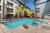 Located in the heart of Scottsdale, Arizona, this outdoor amenity courtyard at 4111 N Drinkwater Blvd offers a resort-style retreat surrounded by a multi-story stucco building with private balconies. The centerpiece is a generously sized swimming pool with crystal-clear turquoise water, complemented by a tiled spa/hot tub positioned conveniently along the pool's edge. A broad concrete pool deck provides ample space for relaxation, lined with rows of blue lounge chairs and shaded umbrellas for sun protection during Arizona's sunny days. Towering palm trees and lush greenery, including bamboo plantings and mature shade trees, create a private, tropical atmosphere within the enclosed courtyard. Raised brick planters add architectural interest and natural texture throughout the space. An outdoor grilling and seating area is tucked alongside the pool deck, offering a functional space for outdoor dining and gatherings. The surrounding multi-story building features wrought iron-railed balconies and a warm desert-toned exterior, framing the courtyard and contributing to the secluded, community-oriented ambiance. Under the abundant Scottsdale sunshine, this well-appointed outdoor space delivers a compelling blend of comfort, leisure, and desert-inspired design.