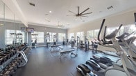 This spacious fitness center is designed to support a full range of workout routines in a bright, welcoming environment. The single-floor layout is thoughtfully organized to maximize usability, with distinct zones for cardio training, strength machines, and free weights. Large multi-pane windows line the walls on multiple sides, flooding the interior with natural light and offering views of the surrounding greenery. Overhead, multiple large ceiling fans provide consistent airflow throughout the space, complementing the recessed lighting and crown molding that add a polished architectural finish. The floors are covered in durable dark rubber matting, ideal for high-impact exercise and heavy equipment use. The cardio section features a row of treadmills and elliptical machines, each equipped with individual monitor screens for entertainment during workouts. The strength training area offers a wide array of cable and selectorized machines targeting various muscle groups, while a full rack of dumbbells lines a mirrored wall, allowing for free weight exercises with easy form checks. Adjustable benches are positioned throughout for versatile use. Wall-mounted televisions provide additional entertainment options across the facility, enhancing the overall workout experience.