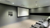 This purpose-built home theater is designed to deliver a true cinematic experience within a private setting. The room is finished with smooth gray walls accented by a bold horizontal black band, lending a sleek, modern aesthetic reminiscent of a professional screening room. A large, centrally mounted projection screen dominates the main wall, providing an expansive viewing surface ideal for movies, sports, and entertainment. Recessed ceiling lights are thoughtfully positioned to allow for controlled, dimmable ambient lighting without detracting from the viewing experience. The ceiling also features a ventilation system to maintain comfort during extended use. Plush leather recliner chairs with built-in cup holders are arranged to face the screen, ensuring optimal sightlines and maximum comfort for viewers. The carpeted flooring adds warmth underfoot while also contributing to the room's acoustic performance. An equipment panel is integrated into the side wall, keeping media components neatly housed and accessible. The overall design prioritizes both function and atmosphere, creating a dedicated space that delivers an immersive, distraction-free entertainment environment.