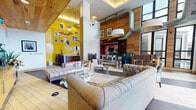 The SkyClub/SkyPark is a rooftop amenity space defined by its striking blend of bold design and open-air living. The interior lounge commands attention with soaring ceilings clad in warm wood paneling, exposed industrial ductwork, and a dramatic yellow accent wall adorned with an eclectic gallery of framed artwork and decorative mirrors. A fireplace serves as a central focal point, flanked by tufted sofas, striped settees, and accent chairs arranged around glass coffee tables, creating a comfortable and stylish gathering environment. A bar area with high stools adds to the social atmosphere, while abundant natural light pours in through large windows.

The kitchen is fully equipped with ample counter space, wood cabinetry, marble-style countertops, and stainless steel appliances beneath an ornate gold pendant fixture—ideal for catered events or communal use.

Expansive folding glass doors open directly onto the outdoor terrace, seamlessly connecting interior and exterior spaces. The terrace features a bold checkerboard tile pattern and is furnished with wicker pod chairs, vibrant turquoise ornate accent chairs, and sunloungers. A covered grill station and planter beds complete the outdoor area, which offers open views of the surrounding city skyline.