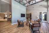 Located at 315 SW Pine Island Rd in Cape Coral, Florida, this single-story commercial space offers a well-designed, contemporary environment ideal for community gatherings, meetings, and recreational activities. The entrance features expansive floor-to-ceiling glass doors and windows that flood the space with abundant natural light and provide views of the surrounding Florida landscape, creating an inviting first impression.

Inside, warm wood-look flooring runs throughout a spacious corridor and lobby area, flanked by sleek glass-panel partitions with dark metal framing that provide transparency while defining separate functional zones. Soaring vaulted ceilings elevate the sense of openness throughout the main hallway and reception areas.

A dedicated conference room offers a professional setting with soft sage-green walls, a large rectangular meeting table with ergonomic seating, a wall-mounted display, and modern pendant lighting overhead — ideal for presentations and collaborative sessions.

The open recreation and lounge area is a standout feature, boasting a bold geometric-patterned floor in teal, orange, and gold tones. A central island with bar-height seating, open shelving, and a mounted television serves as a social hub, while a shuffleboard table adds a recreational element. The space is completed with stylish pendant lighting and easy access to the exterior, making it a vibrant and functional gathering destination.