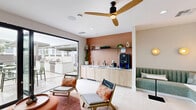 Located in Phoenix, AZ, this contemporary single-floor lounge space offers a thoughtfully designed living area that seamlessly blends indoor comfort with outdoor access. The interior is anchored by a striking floor-to-ceiling living moss wall, adding a vibrant, natural focal point to the bright, white-walled room. Light-toned porcelain tile flooring runs throughout, enhancing the clean, modern aesthetic.

The lounge features a variety of seating arrangements, including a built-in tufted banquette with attached pedestal tables, woven egg chairs, a neutral sofa with accent pillows, and colorful ottoman seating — all curated to encourage social interaction. Warm globe wall sconces and a sleek dual-tone ceiling fan provide both functional lighting and stylish detail.

A built-in wet bar along one wall is finished with warm wood cabinetry and a terracotta-toned vertical tile backsplash, complete with open shelving for plants and décor, a sink, and a coffee station. A large sliding glass door opens directly to an expansive outdoor terrace, where white marble-top dining tables with sage green chairs are arranged alongside a tranquil water feature wall and a curated lounge seating area with potted olive trees. A grill station is also accessible from the terrace, making this space well-suited for both relaxation and entertaining.
