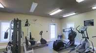 Located at 1000 Commons Circle in Vicksburg, Mississippi, this fitness center offers a practical and functional space designed to support a well-rounded exercise routine. The single-floor facility features a clean, straightforward layout with light-toned walls and a textured ceiling fitted with bright fluorescent lighting, ensuring a well-illuminated environment for workouts at any time of day. Natural light also filters in through a windowed door and a side window, adding a welcoming sense of openness to the space. The room is equipped with a multi-station cable weight machine for strength training, a stationary upright bicycle, an elliptical trainer, and a stair climber, providing a comprehensive range of cardiovascular and resistance exercise options. Rubber floor mats are positioned beneath the equipment to provide stability and protect the flooring. The space is kept accessible with a clearly marked exit door and a fire safety pull station on the wall. The overall ambiance is utilitarian and focused, making it a convenient and straightforward fitness resource for those in the surrounding community.