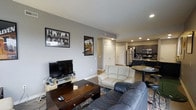 This single-floor residence features two bedrooms and two bathrooms within a generously sized floor plan. The open-concept living and kitchen area serves as the heart of the home, with recessed lighting illuminating the space throughout. The living room comfortably accommodates multiple seating options, as seen with the combination of a leather sofa and a light-colored loveseat arranged around a dark wood coffee table, with ample room for a media console and flat-screen television. Gray-toned walls provide a neutral backdrop that complements the modern aesthetic. The kitchen is visible from the living area and features dark cabinetry, stainless steel appliances including a refrigerator, microwave, and range, as well as a dark countertop breakfast bar with seating. The seamless flow between the living and kitchen areas makes the space well-suited for both everyday living and entertaining. A closet with white bifold doors is visible in the background, adding practical storage. The recessed lighting and open sightlines throughout contribute to a bright, cohesive atmosphere across the entire layout.
