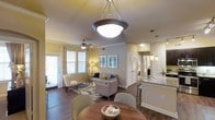 This single-floor residence showcases a thoughtfully designed open-concept floor plan that effortlessly connects the dining, living, and kitchen areas into one cohesive living space. Rich hardwood-style flooring runs throughout, anchoring the warm and inviting atmosphere.

The dining area is highlighted by a striking bowl pendant light fixture that serves as a focal point, complemented by a round dining table and stylish chairs. Adjacent to it, the living room is generously proportioned, featuring a ceiling fan, large windows dressed with elegant drapes, and abundant natural light that fills the space. The seating arrangement, centered on a patterned area rug, creates a comfortable and welcoming environment.

The kitchen is visible from the main living area and features white cabinetry, granite or stone-look countertops, pendant lighting over a breakfast bar with seating, and stainless steel appliances. Crown molding and neutral warm-toned walls add a refined, polished touch throughout the space.

Closet access and additional rooms are visible toward the rear of the unit, suggesting well-organized storage and private spaces. Overall, the residence balances functionality and aesthetics, offering a bright, open, and elegantly appointed living environment ideal for both everyday living and entertaining.