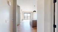 Located at 200 N Adams St in downtown Green Bay, Wisconsin, this modern one-bedroom, one-bathroom unit offers a thoughtfully designed layout across a single floor. The open-concept kitchen serves as the centerpiece of the living space, anchored by a large island with a granite countertop, an undermount sink, and warm wood cabinetry. Stainless steel appliances — including a refrigerator, range, and built-in microwave — complement the crisp white upper cabinets and subway tile backsplash. Two statement dome pendant lights hang above the island, adding a contemporary touch. Light hardwood flooring flows throughout the main living areas, creating a bright and cohesive aesthetic. The bedroom is carpeted for comfort and features a large black-framed window that frames views of the downtown Green Bay skyline, allowing abundant natural light to fill the room. An adjoining walk-in closet provides generous storage with built-in shelving and hanging rods. The bathroom is finished with tile flooring, a granite-topped wood vanity, and a glass-enclosed walk-in shower. A dedicated laundry area with a stacked washer and dryer unit adds everyday convenience. This well-appointed unit combines modern finishes with functional design in the heart of downtown Green Bay.
