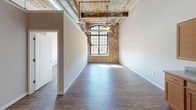 This single-floor unit showcases a compelling blend of industrial character and modern functionality. The open layout is anchored by soaring ceilings with exposed wooden beams and visible ductwork, lending an authentic loft aesthetic throughout. Dark hardwood floors run continuously across the living areas, creating a warm contrast against the raw, exposed brick walls that serve as a defining architectural feature.

Large, multi-pane industrial-style windows allow generous amounts of natural light to pour into the living spaces, framing views of the surrounding area while highlighting the textural richness of the brick. The arched detailing on select windows adds a refined architectural touch to the otherwise industrial character of the unit.

The kitchen is well-appointed with warm-toned wood cabinetry, granite-style countertops, and a full suite of stainless steel appliances, including a dishwasher, range, microwave, and refrigerator. Pendant lighting above the kitchen area complements the overall aesthetic.

The bathroom includes a dedicated walk-in shower area with tile flooring, a towel bar, and neutral finishes, offering a clean and functional design. The 3D floorplan reveals a spacious, well-organized layout with distinct zones for living, dining, and sleeping, all benefiting from the unit's impressive ceiling height.
