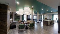 This single-floor communal space is thoughtfully designed to foster social interaction while maintaining a sophisticated aesthetic. The most striking architectural feature is the bold teal-painted ceiling, which adds a vibrant pop of color and creates a visually dynamic atmosphere throughout the open-plan layout. Large drum-style pendant chandeliers hang in clusters, providing warm, ambient illumination that complements the recessed ceiling lights throughout the space.

The lounge area is furnished with a curated mix of seating arrangements, including patterned green accent chairs, a neutral sofa, and sleek coffee tables — all positioned to encourage conversation and relaxation. The wide-plank wood-look flooring in cool grey tones runs seamlessly across the entire floor, unifying the various zones.

Structural columns are integrated throughout, subtly defining different areas without disrupting the open feel. Toward the rear of the space, a billiards table serves as a central recreational focal point. Adjacent to this, a bar-style counter with pendant lighting and bar stools provides a casual dining or socializing area. Large windows and glass doors along the perimeter allow abundant natural light to flood the interior, enhancing the overall brightness and airy ambiance of the space.