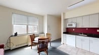 Located at 327 Eddy St in Ithaca, NY, this three-bedroom, one-bathroom unit offers a practical and functional layout across a single floor. The combined kitchen, dining, and living area forms the heart of the space. The kitchen is well-equipped with gray cabinetry extending to the ceiling, dark granite-style countertops, a bold red tile backsplash, white appliances including a stove, refrigerator, dishwasher, and microwave, and tile flooring. Adjacent to the kitchen, the dining area accommodates a round wooden table with chairs and is brightened by a window with views of the surrounding neighborhood. The compact living room features blue-gray carpeting and a teal entry door, adding a pop of color to the neutral palette. Each of the three bedrooms is carpeted and includes a dedicated closet and dresser for storage. The primary bedroom stands out with corner windows that frame sweeping panoramic views of Ithaca's rolling hills and cityscape, filling the room with natural light. Additional bedrooms are similarly functional, featuring glass-block windows for privacy and natural light. The shared bathroom includes tile flooring, a vanity with a speckled countertop, and a mirrored medicine cabinet. Throughout the unit, cream-colored walls and blue-gray carpet create a cohesive, understated aesthetic.