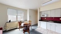 Located at 327 Eddy St in Ithaca, NY, this three-bedroom, one-bathroom unit offers a practical and functional layout across a single floor. The combined kitchen, dining, and living area forms the heart of the space. The kitchen is well-equipped with gray cabinetry extending to the ceiling, dark granite-style countertops, a bold red tile backsplash, white appliances including a stove, refrigerator, dishwasher, and microwave, and tile flooring. Adjacent to the kitchen, the dining area accommodates a round wooden table with chairs and is brightened by a window with views of the surrounding neighborhood. The compact living room features blue-gray carpeting and a teal entry door, adding a pop of color to the neutral palette. Each of the three bedrooms is carpeted and includes a dedicated closet and dresser for storage. The primary bedroom stands out with corner windows that frame sweeping panoramic views of Ithaca's rolling hills and cityscape, filling the room with natural light. Additional bedrooms are similarly functional, featuring glass-block windows for privacy and natural light. The shared bathroom includes tile flooring, a vanity with a speckled countertop, and a mirrored medicine cabinet. Throughout the unit, cream-colored walls and blue-gray carpet create a cohesive, understated aesthetic.