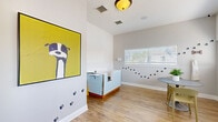 Located at 100 Reflections Cir in Casselberry, Florida, this single-room office space is thoughtfully designed to serve as a professional pet care environment. The interior features warm, light wood-tone flooring that runs throughout, paired with neutral gray walls that create a clean and calming backdrop. The space is enlivened with whimsical dog-themed décor, including paw print wall decals trailing across the walls, large photographic dog portraits displayed near the windows, and a bold, colorful canine artwork that serves as a striking focal point. Multiple windows dressed with white horizontal blinds allow generous natural light to fill the room, contributing to an open and welcoming atmosphere. A light blue grooming or bathing station is positioned prominently within the space, offering a functional centerpiece for pet care services. A round marble-top table with seating provides a consultation or waiting area, while a stainless steel utility table offers additional workspace. The vaulted ceiling adds a sense of height and openness beyond the room's footprint. A glass-paned door provides access to an adjoining area. Overall, the space strikes a balance between professional functionality and a warm, pet-friendly ambiance that is sure to appeal to animal owners.
