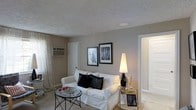 This single-floor space presents a thoughtfully arranged living area finished in warm neutral tones that create a calm, welcoming atmosphere. The living room serves as the central gathering space, featuring a plush white sofa accented with black decorative pillows, which provides a clean contrast against the taupe-painted walls. A textured popcorn ceiling adds a classic architectural character overhead, while a flush-mount light fixture provides ambient illumination.

Two framed black-and-white photographs are displayed side by side on the main wall, contributing an artistic focal point to the room. A tall, cone-shaped floor lamp positioned beside a sleek glass-and-metal side table adds both functional lighting and decorative interest. A round glass-top coffee table with a slender black metal base anchors the seating area.

Natural light enters through a window dressed with sheer white curtains, enhanced by a wall-mounted air conditioning unit. A doorway visible on the right suggests additional rooms beyond, offering a sense of continued living space. The carpeted flooring ties the room together, completing the comfortable and cohesive aesthetic of this well-appointed interior.