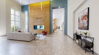 Located at 899 N Orange Ave in the heart of Orlando, Florida, this expansive single-floor amenity lounge offers a generously sized, open-plan gathering space designed with a contemporary aesthetic. Soaring double-height ceilings amplify the sense of openness throughout, while large floor-to-ceiling windows flood the interior with abundant natural light and offer views of the surrounding outdoor areas.

The space is anchored by a dramatic floor-to-ceiling wood-paneled feature wall, adorned with sculptural metalwork and a built-in pass-through window that connects to an adjacent service area. Multiple distinct seating zones are thoughtfully arranged throughout, including plush white sofas, bold terracotta accent chairs, and intimate conversational groupings, all set atop area rugs over neutral large-format tile flooring.

A standout entertainment zone features a shuffleboard table, a large multi-panel TV display, and a vivid botanical-themed mural celebrating Orlando's citrus heritage. Additional areas include a dining table with modern seating, a rustic wood shelving unit displaying curated décor, and a lounge area anchored by a striking abstract wall mural. The overall ambiance balances vibrant color accents — teal, gold, and terracotta — with clean white walls and warm wood tones, creating an inviting, energetic communal environment.
