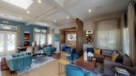 This well-appointed single-floor community clubhouse offers an open, flowing layout designed for both relaxation and socializing. The main lounge area is generously sized, featuring light hardwood flooring, coffered ceilings with recessed lighting, and large windows dressed with sheer curtains and wood-slat blinds that allow abundant natural light to fill the space. Seating is arranged thoughtfully throughout, including plush teal velvet accent chairs with gold-toned legs, a taupe sofa accented with navy and mustard throw pillows, and a warm walnut coffee table — all anchored by a patterned area rug.

A standout architectural feature is the wood-paneled partition that frames a private dining nook, complete with built-in upholstered bench seating, a pedestal table, and a woven rattan pendant light overhead. An adjacent dining area with a long communal table and modern chairs sits beneath a teal accent wall adorned with contemporary artwork.

The space also includes a kitchenette area highlighted by a striking white marble waterfall island with bar seating, a built-in microwave and beverage station, a wall-mounted TV framed by a stone tile accent wall, and a textured metallic ceiling detail. French doors lead to an additional game room beyond. Large glazed entry doors open to outdoor views, completing this vibrant and versatile community gathering space.