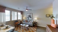 This single-floor living space features a thoughtfully designed living room that balances comfort and contemporary style. The room is finished with hardwood-style flooring that adds warmth and visual continuity throughout the space. A textured ceiling with a ceiling-mounted fan provides both airflow and a subtle architectural detail overhead. Natural light enters through a window dressed with horizontal blinds and patterned curtains, brightening the interior and creating an inviting atmosphere. The neutral wall tones serve as a versatile backdrop, complemented by a bold abstract artwork that serves as a focal point above the seating area. The living area comfortably accommodates a sofa with accent pillows, a mid-century modern accent chair, and a rectangular coffee table set atop a geometric-patterned area rug in warm orange and cream tones. Side tables with table lamps on either side of the sofa provide balanced ambient lighting. The overall ambiance is modern yet approachable, combining clean lines with warm textures and curated décor to create a cohesive and livable environment well-suited for everyday use and relaxed entertaining.