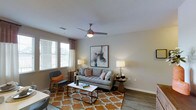 This single-floor living space features a thoughtfully designed living room that balances comfort and contemporary style. The room is finished with hardwood-style flooring that adds warmth and visual continuity throughout the space. A textured ceiling with a ceiling-mounted fan provides both airflow and a subtle architectural detail overhead. Natural light enters through a window dressed with horizontal blinds and patterned curtains, brightening the interior and creating an inviting atmosphere. The neutral wall tones serve as a versatile backdrop, complemented by a bold abstract artwork that serves as a focal point above the seating area. The living area comfortably accommodates a sofa with accent pillows, a mid-century modern accent chair, and a rectangular coffee table set atop a geometric-patterned area rug in warm orange and cream tones. Side tables with table lamps on either side of the sofa provide balanced ambient lighting. The overall ambiance is modern yet approachable, combining clean lines with warm textures and curated décor to create a cohesive and livable environment well-suited for everyday use and relaxed entertaining.