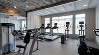 The fitness center at 7 N Main is a thoughtfully designed single-floor space that balances contemporary functionality with distinctive architectural character. The ceiling is one of the room's most striking features, combining rustic recessed wood planking with white-painted structural beams, creating a compelling contrast that adds warmth and texture to the otherwise modern environment.

Large windows line the perimeter wall, allowing abundant natural light to fill the space and offering views of the surrounding exterior. White horizontal blinds provide flexible light control while maintaining the room's bright, open atmosphere. The generous floor-to-ceiling height contributes to an airy, spacious feel throughout.

The fitness area is outfitted with a well-curated selection of cardio equipment, including multiple treadmills, an elliptical trainer, and a stationary cycling machine. Full-length mirrored walls along one side visually expand the space and provide practical utility for monitoring form during workouts. Recessed ceiling lighting supplements the natural light, ensuring consistent illumination. The neutral, carpeted flooring ties the aesthetic together, creating a clean, professional fitness environment.
