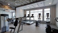 The fitness center at 7 N Main is a thoughtfully designed single-floor space that balances contemporary functionality with distinctive architectural character. The ceiling is one of the room's most striking features, combining rustic recessed wood planking with white-painted structural beams, creating a compelling contrast that adds warmth and texture to the otherwise modern environment.

Large windows line the perimeter wall, allowing abundant natural light to fill the space and offering views of the surrounding exterior. White horizontal blinds provide flexible light control while maintaining the room's bright, open atmosphere. The generous floor-to-ceiling height contributes to an airy, spacious feel throughout.

The fitness area is outfitted with a well-curated selection of cardio equipment, including multiple treadmills, an elliptical trainer, and a stationary cycling machine. Full-length mirrored walls along one side visually expand the space and provide practical utility for monitoring form during workouts. Recessed ceiling lighting supplements the natural light, ensuring consistent illumination. The neutral, carpeted flooring ties the aesthetic together, creating a clean, professional fitness environment.