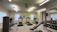 This single-floor fitness center offers a functional and well-organized workout environment designed to accommodate a variety of exercise routines. The space is outfitted with multiple treadmills featuring integrated display screens, an elliptical trainer, a stationary bike, and a flat workout bench, providing a thorough range of cardio and strength training options. A stability ball and additional equipment round out the available fitness tools.

The room features light-toned, patterned carpet flooring that adds a degree of comfort underfoot. Overhead, a drop tile ceiling is illuminated by several fluorescent strip lights and a ceiling fan, ensuring the space remains well-lit and ventilated during use. Natural light filters in through multiple windows fitted with white blinds, contributing to an open and airy atmosphere.

The walls are painted in a neutral, light tone, keeping the environment calm and focused. Framed artwork adorns one wall, adding a subtle decorative touch. A large mirror on one wall enhances the sense of space and allows users to monitor their form. The overall ambiance is clean, practical, and purposefully arranged to maximize usability and comfort for fitness activities.