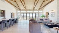 This single-story community space is a masterclass in contemporary design, blending warm natural materials with an open, airy layout. The centerpiece of the main gathering area is a dramatic vaulted ceiling adorned with exposed rustic wood beams over a light-toned shiplap surface, creating a striking architectural contrast against the crisp white walls. Light wide-plank hardwood flooring extends throughout, unifying the space with warmth and continuity.

The main lounge area is generously proportioned, anchored by a large sectional sofa, rounded upholstered accent chairs, and a full-height stone fireplace with an integrated media wall. Floor-to-ceiling black-framed sliding glass doors and transom windows line the rear wall, flooding the interior with natural light and offering direct views and access to an outdoor amenity area.

The entry foyer is welcoming and light-filled, with tall windows framing outdoor views and a small seating arrangement for informal gatherings. Adjacent to the main lounge, a bar-height island with seating is positioned alongside built-in shelving stocked with amenities, functioning as a café-style service area. A separate reading nook or flex room features built-in bookshelves, a sliding barn door, and additional natural light. Steel-framed glass interior doors provide visual connectivity between zones while maintaining a refined, modern aesthetic throughout.