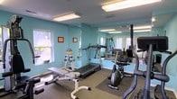 Located at 1192 Harbour Dr in Wilmington, NC, this fitness center is a compact yet well-equipped single-floor facility designed to support a well-rounded workout routine. The space is finished with light teal walls that create a clean, energizing atmosphere, complemented by bright overhead fluorescent lighting and natural light streaming in through multiple windows. A large wall-mounted mirror expands the sense of space and allows for proper form checks during exercise. The facility is equipped with a treadmill, a recumbent stationary bike, and a multi-station cable strength machine, catering to both cardio and resistance training needs. A dedicated free weight area includes a dumbbell rack with a range of weights alongside an adjustable workout bench, offering versatility for strength training. The flooring features a combination of carpet and protective rubber mats beneath the equipment, providing both comfort and safety. Motivational framed prints add a personal touch to the walls. The space is thoughtfully organized to maximize usability while maintaining clear pathways throughout, making it a functional and welcoming environment for daily exercise.