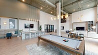 Located at 700 SE Veranda Pl in Port Saint Lucie, Florida, this thoughtfully designed community living space offers an expansive, open-concept layout that seamlessly blends functionality with contemporary style. The single-floor space is defined by its dramatic soaring ceilings, which are accented by exposed industrial elements and decorative pendant lighting that draw the eye upward and amplify the sense of openness throughout.

Light wood-look flooring extends across the entire space, complementing the neutral, modern color palette of whites, grays, and sage greens. Oversized black-framed windows and glass doors flood the interior with natural light while offering views of the lush Florida landscape, including swaying palm trees beyond the exterior.

The living area features a bold accent wall with a textured stone fireplace surround, built-in shelving, and a mounted television, serving as a striking focal point. A generous sectional seating arrangement creates a comfortable and inviting gathering space. A game area with a pool table adds a recreational element, while a banquette dining nook with tufted bench seating and café-style tables provides a cozy spot for socializing. A granite-topped island and a wet bar with mint-green cabinetry offer stylish, practical amenities. The space is completed by a casual bar-height dining area near the entrance, rounding out this well-appointed community retreat.