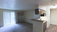 This single-floor unit features an open-concept design that integrates the living area and kitchen into one cohesive space. The living area is finished with wall-to-wall carpeting and neutral-toned walls, creating a clean, functional environment. A built-in shelving unit is positioned adjacent to a sliding glass door, which allows natural light to fill the space and provides direct access to an outdoor area. The textured ceiling adds a classic touch throughout.

The kitchen is positioned along one side of the open layout and features a peninsula counter with a dark laminate surface, providing both workspace and a natural divider between the kitchen and living areas. Light wood cabinetry lines the upper walls, offering ample storage, while a range and sink are integrated into the countertop. An overhead cabinet extends above the peninsula, adding additional storage space. A doorway visible from the main area leads to additional rooms within the unit. Overall, the layout maximizes functional use of space with a practical, straightforward design.