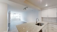 This single-floor space showcases a clean, modern open-concept layout that integrates the kitchen and living area into one cohesive environment. The kitchen is anchored by a generous granite-topped island with a stainless steel undermount sink and a contemporary pull-down faucet, offering both functional workspace and a natural gathering point. White subway tile backsplash lines the kitchen walls, complementing the white cabinetry above for a crisp, cohesive aesthetic.

Track lighting overhead provides focused illumination across the space, while the living area benefits from a large window that allows natural light to pour in and frames views of the surrounding urban streetscape. The light-toned hardwood-style flooring runs throughout, unifying the open layout and contributing to an airy, expansive feel. Neutral white walls and a clean ceiling with subtle structural beams reinforce the contemporary character of the space.

Overall, the unit presents a minimalist, well-lit environment well-suited to those seeking a modern urban residence with practical, stylish finishes and an open, flowing floor plan.