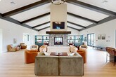 Located at 1 Ridge Rd in Auburn, Massachusetts, this single-story community clubhouse offers a generously sized, open-concept layout designed for both relaxation and social engagement. The space is defined by its dramatic vaulted ceilings adorned with dark exposed beams, which lend a modern farmhouse character throughout. Light hardwood flooring runs continuously across the entire floor, enhancing the bright, airy atmosphere created by abundant natural light pouring in through large, multi-pane black-framed windows.

The central gathering area serves as the heart of the clubhouse, anchored by a striking stone fireplace with a substantial wood mantel and a wall-mounted television above. Teal built-in shelving flanks the fireplace, adding a pop of color and functional display space. Multiple seating arrangements with a mix of plush sofas and accent chairs invite casual conversation and comfortable lounging. A statement chandelier adds warmth and visual interest overhead.

Adjacent to the main lounge, a game area features a billiards table, while a separate lounge with floor-to-ceiling windows offers scenic outdoor views. A kitchenette with teal cabinetry provides convenient amenities. A private conference room with a wall-mounted display and a decorative area rug offers a quieter setting for meetings or focused gatherings. The entrance and hallway connect all areas seamlessly, completing this well-appointed communal space.