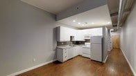 This single-floor space showcases a well-appointed kitchen that serves as a central feature of the layout. The kitchen is outfitted with white cabinetry spanning both upper and lower configurations, providing ample storage. A dark mosaic tile backsplash adds a contrasting design element against the white cabinetry and light countertops. White appliances, including a dishwasher and range, complement the kitchen's clean aesthetic, while a stainless steel refrigerator introduces a modern accent. Recessed lighting illuminates the kitchen area effectively, enhancing the overall brightness of the space. The kitchen transitions seamlessly into an open adjacent living area, both unified by warm hardwood-style flooring that runs throughout. The walls are finished in a neutral gray tone, creating a clean and contemporary backdrop. Generous baseboard trim lines the perimeter, adding a polished architectural detail. The open layout between the kitchen and living area creates a sense of spaciousness, making the space feel practical and inviting for everyday living.