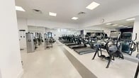 Located at 3 Davenport Avenue in New Rochelle, NY, this fitness center offers a thoughtfully organized single-level layout designed to support a full range of workout routines. The facility is divided into distinct functional zones, each tailored to a specific type of training.

The main cardio area features a row of treadmills and elliptical machines arranged along a wall of large, floor-to-ceiling mirrors that visually expand the space. The terrazzo flooring, accented with neutral striping, lends a clean and professional aesthetic throughout. Recessed panel lighting illuminates the space evenly, while a wall clock keeps members on schedule.

Adjacent to the cardio zone is a dedicated strength training area, distinguished by its vibrant blue rubber flooring and deep plum accent walls. This room is equipped with a multi-station cable machine, a dumbbell rack with a full range of free weights, and wall-mounted resistance training attachments. Mirrors line the walls here as well, allowing for proper form monitoring during lifts.

The entry area provides a welcoming transition into the gym, featuring glass double doors, a wall mirror, and Cybex stationary bikes positioned near the entrance. Stability balls are neatly stored nearby, adding to the facility's versatility.

An accessible restroom completes the layout, finished with large-format marble-look tile, grab bars, and a clean white palette. The gym offers a practical, modern environment suited for focused fitness training.