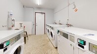 This shared laundry facility is a well-organized, single-room space designed for communal use. The room is finished with durable terrazzo flooring throughout, providing a practical and easy-to-clean surface suited to high-traffic use. White-painted walls and a flat ceiling fitted with fluorescent tube lighting ensure the space is bright and functional at all times.

The facility is equipped with a row of commercial-grade front-load washing machines and matching dryers, all in white, arranged along the walls to maximize available floor space. Each machine features a digital control panel, indicating modern, programmable operation. Plumbing connections with clearly visible water taps are positioned along the wall behind the machines. A top-load washing machine is also present, offering users additional flexibility.

A wall-mounted dispenser unit provides convenient access to laundry products. Seating is available via a plastic chair, accommodating users during wait times. A dedicated wall panel displays notices and usage instructions for residents.

The room is accessed via a solid timber-framed door and includes a wall-mounted ventilation unit to maintain air circulation. A smoke detector is also visible on the ceiling, reflecting attention to safety. The overall layout is clean, efficiently arranged, and well-maintained, making it a practical shared amenity.