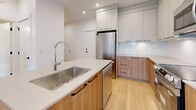 This well-appointed one-bedroom, one-bathroom unit at 15624 24 Ave in Surrey, BC offers a thoughtfully designed layout across a single floor. Light hardwood flooring flows throughout, creating a warm, cohesive aesthetic from room to room.

The kitchen is a standout feature, showcasing a central island with a quartz countertop and an undermount stainless steel sink, warm wood cabinetry paired with soft grey upper cabinets, stainless steel appliances including a full-size refrigerator, microwave, and range, and recessed lighting that brightens the entire space. Ample upper and lower cabinetry provides generous storage.

The bedroom is a calm, airy retreat with light-toned hardwood floors, neutral walls, and a large picture window that frames views of mature trees and the surrounding neighbourhood, allowing natural light to fill the room.

The bathroom is finished in a clean, modern style with large-format floor tiles, a floating wood vanity with a white quartz countertop and undermount sink, black hardware accents, and a glass-enclosed shower stall. A hallway connects the rooms efficiently, completing the functional floor plan.

Overall, the unit presents a contemporary and cohesive design with quality finishes throughout.