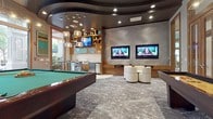 This sophisticated entertainment lounge is designed with a strong contemporary aesthetic, combining sleek architectural details with a vibrant, social atmosphere. The space is defined by a dramatic curved, dark ceiling with recessed lighting and layered tiers that add depth and visual interest overhead. Clusters of globe pendant lights hang above the bar area, casting a warm, ambient glow throughout.

The room is thoughtfully divided into distinct activity zones. A full-size pool table anchors the central area, complemented by a shuffleboard table along one side. Adjacent to the gaming area, a wet bar station features warm wood cabinetry with open shelving, a sink, under-counter refrigeration, and high-top tables with chrome barstools, ideal for socializing.

A dedicated media lounge area showcases a textured wave-patterned accent wall hosting dual flat-screen televisions, with curved white lounge chairs arranged for comfortable viewing. Additional wall-mounted screens are integrated throughout. Large wood-framed glass doors and windows on multiple walls flood the space with natural light and offer views of an outdoor pool area and palm trees, seamlessly connecting the interior to the surrounding environment. The flooring features richly patterned stone-effect carpeting throughout.