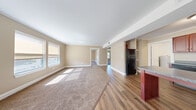 This single-floor space is designed with a practical, open layout that connects the main living area to the kitchen and adjoining rooms. The living area is generously sized, featuring warm beige walls, white trim, and plush neutral-toned carpet that transitions seamlessly into wood-style laminate flooring toward the kitchen and hallway. Multiple windows allow natural light to fill the space, casting bright patches across the floors and creating a welcoming atmosphere.

The kitchen is equipped with rich, dark cherry-toned wood cabinetry offering ample upper and lower storage, granite-style countertops, a black refrigerator, a black dishwasher, and a double sink. A kitchen island with a dark countertop adds additional workspace. An adjacent utility area houses a water heater, providing added convenience.

The bedrooms are carpeted and finished with the same warm neutral palette, crown molding, and white baseboards. Each room features windows that bring in natural daylight and views of the surrounding area, along with closet space for storage.

Throughout the unit, consistent design elements — including neutral wall tones, clean white trim, and a cohesive flooring scheme — contribute to a tidy, move-in-ready feel.