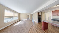 This single-floor space is designed with a practical, open layout that connects the main living area to the kitchen and adjoining rooms. The living area is generously sized, featuring warm beige walls, white trim, and plush neutral-toned carpet that transitions seamlessly into wood-style laminate flooring toward the kitchen and hallway. Multiple windows allow natural light to fill the space, casting bright patches across the floors and creating a welcoming atmosphere.

The kitchen is equipped with rich, dark cherry-toned wood cabinetry offering ample upper and lower storage, granite-style countertops, a black refrigerator, a black dishwasher, and a double sink. A kitchen island with a dark countertop adds additional workspace. An adjacent utility area houses a water heater, providing added convenience.

The bedrooms are carpeted and finished with the same warm neutral palette, crown molding, and white baseboards. Each room features windows that bring in natural daylight and views of the surrounding area, along with closet space for storage.

Throughout the unit, consistent design elements — including neutral wall tones, clean white trim, and a cohesive flooring scheme — contribute to a tidy, move-in-ready feel.