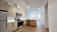 Located at 808 Chapel Street in the heart of New Haven, Connecticut, this thoughtfully designed single-floor unit offers a modern, efficient layout that maximizes every square foot. The open-concept kitchen and living area serves as the centerpiece of the space, featuring light wood cabinetry that extends to the ceiling, sleek white quartz countertops, and a full suite of stainless steel appliances including a refrigerator, electric range, built-in microwave, and dishwasher. Track lighting overhead highlights the clean lines of the kitchen, which flows seamlessly into the bright living area. A window frames views of the surrounding urban streetscape, allowing natural light to fill the space. Wood-look flooring runs throughout, tying each room together with warmth and continuity. The bathroom is finished with matching light wood cabinetry, a white quartz vanity, a large mirror, and a glass-enclosed shower. A dedicated walk-in closet with built-in wire shelving provides generous storage. The layout is completed by a hallway that connects all rooms efficiently. Neutral white walls and consistent modern finishes throughout give the space a clean, contemporary ambiance well-suited to urban living in New Haven.
