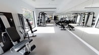 This fitness center is housed in a single-level, elongated space designed to accommodate a comprehensive range of exercise equipment and training activities. The layout is thoughtfully organized, with equipment arranged to maximize floor space and allow for easy movement throughout the facility.

The interior is finished with a clean, light-toned rubber flooring that provides both durability and comfort underfoot. Full-length mirrors line one wall, enhancing the sense of space while allowing users to monitor their form during workouts. Large floor-to-ceiling windows along the opposite wall flood the space with abundant natural light and offer views of the surrounding greenery, creating an inviting and energizing atmosphere.

The strength training section is well-appointed with cable machines, multi-functional weight stations, adjustable benches, and free weight equipment. The cardio zone features a row of treadmills and additional aerobic machines, positioned to take advantage of the natural light from the windows. Overhead, recessed linear lighting and clean white ceilings contribute to the bright, modern aesthetic.

The overall ambiance is sleek and professional, with a monochromatic black-and-white color scheme throughout the equipment and finishes. The space is designed to serve a wide range of fitness needs, from strength training to cardiovascular conditioning, in a clean and well-maintained environment.