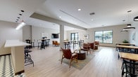 This well-appointed single-floor communal space is designed to accommodate a variety of social and functional needs, blending modern aesthetics with practical versatility. The open-plan layout is anchored by light wood-look tile flooring and neutral gray walls, creating a clean, welcoming atmosphere throughout.

The space is thoughtfully divided into several distinct zones. A casual lounge area features mid-century-inspired leather armchairs arranged around a cylindrical wood coffee table atop a patterned area rug, accompanied by a wall-mounted flat-screen TV and a bold circular pendant light overhead. Adjacent to this is a dining section with natural wood-top tables and black wire chairs, illuminated by matching circular pendant fixtures and accented by vibrant framed artwork on the walls.

A dedicated bar and kitchen area includes a large white quartz countertop with subway tile detailing, adjustable industrial-style bar stools, stainless steel appliances, and a wall-mounted TV — ideal for casual entertaining. Additional high-top bar tables with black stools provide a more informal seating option.

Large windows along one wall flood the interior with natural light and offer views to the exterior. Curated contemporary artwork is displayed throughout, reinforcing the polished, modern character of the space.