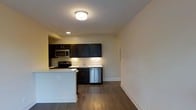 This single-floor space showcases a thoughtfully designed open-concept layout that seamlessly connects the kitchen and living area. The kitchen is a standout feature, equipped with rich, dark espresso-toned cabinetry that provides ample storage both above and below the countertops. Stainless steel appliances, including a range, built-in microwave, and dishwasher, lend a modern, polished aesthetic. The light-colored countertops contrast sharply with the dark cabinetry, creating a visually appealing balance. A peninsula counter extends into the living area, offering additional workspace or casual dining options.

The flooring throughout is a warm hardwood-style material that adds continuity and a sense of warmth to the open space. The walls are finished in a clean, neutral tone that enhances the brightness of the interior. Overhead, flush-mounted ceiling lighting provides consistent illumination across both the kitchen and adjoining living area. Recessed lighting above the kitchen adds focused task lighting. The overall ambiance is clean, contemporary, and functional, making efficient use of the available space.