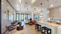 This single-story clubhouse offers a thoughtfully designed community space that blends contemporary aesthetics with functional versatility. Light wood-look flooring runs throughout, tying together the various zones and creating a warm, cohesive atmosphere.

The main lounge area is anchored by a dramatic floor-to-ceiling accent wall finished in a veined stone-look material with gold detailing, displaying curated artwork and sconce lighting. Seating clusters featuring upholstered chairs, organic wood stump tables, and area rugs invite casual socializing. Large black-framed glass doors flood the space with natural light and provide direct outdoor access.

An open bar-height counter with bar stools serves as a social hub adjacent to the full catering kitchen, which is outfitted with light wood cabinetry, quartz countertops, a tiled backsplash, a sink, and under-counter appliances. A dark wood slatted partition elegantly separates kitchen zones while maintaining visual openness.

A quieter seating lounge features the same stone accent wall alongside teal-painted walls, upholstered chairs, and gold-accented side tables for more intimate gatherings. A private dining nook with floor-to-ceiling windows offers garden views. A separate work or service counter area with antique mirror backsplash completes the layout.