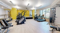 Located at 3240 Champion Ring Rd in Fort Myers, Florida, this fitness center offers a generously sized, single-floor layout designed to accommodate a full range of workout activities. The space is divided into two distinct zones — a cardio area and a strength training area — connected by a railed transition ramp, giving the facility a dynamic, multi-level feel within a single floor.

The cardio zone features light wood-look flooring and is lined with large windows that flood the area with natural light and provide outdoor views of lush surroundings. Bold yellow accent walls energize the space, complemented by a ceiling fan and linear LED lighting overhead. Matrix-brand equipment, including stair climbers, treadmills, and upright bikes, is thoughtfully arranged to maximize usability and floor space.

The strength training area transitions to neutral tones with white walls and continues the light flooring aesthetic. Full-length mirrors expand the visual depth of the room, while cable machines, weight benches, and functional training stations provide comprehensive options for resistance training. Recessed lighting and a ceiling fan maintain comfort throughout.

Together, the two zones create a bright, modern, and well-appointed fitness facility suited for a wide variety of exercise routines.
