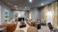 This thoughtfully designed single-floor community space combines contemporary style with a warm, inviting atmosphere. The main lounge area serves as the centerpiece of the space, featuring rich cognac leather sofas arranged around a dark wood coffee table atop a patterned area rug. Woven pouf ottomans add casual seating and texture, while recessed lighting illuminates the grey-toned walls adorned with an eclectic cluster of decorative plates and a large abstract canvas. A dark credenza anchors one wall, styled with table lamps and accent décor in warm yellow tones that carry throughout the space via bold, geometric-print drapery panels. Adjacent to the lounge, a semi-private dining nook offers a round marble-top table with sleek charcoal leather chairs, ideal for small meetings or casual gatherings. A vibrant abstract painting in blue and gold tones decorates the wall, while floor-length yellow patterned curtains provide a cohesive design thread. Large glass partitions connect the lounge to a visible fitness center beyond, bringing in additional light and a sense of openness. The overall palette of warm greys, cognac, and golden yellow creates a polished yet comfortable environment throughout.