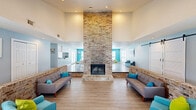 Located in Sherwood, Arkansas, this single-floor community clubhouse offers a well-designed gathering space that blends rustic charm with modern, colorful décor. The centerpiece of the living area is a striking floor-to-ceiling brick fireplace that anchors the room and draws the eye upward toward soaring ceilings, creating an impressive sense of vertical space. Low brick half-walls define the sunken seating areas on either side of the fireplace, where contemporary gray sofas with teal accent pillows provide comfortable lounge seating atop light-toned hardwood flooring. A sliding barn door adds a stylish architectural detail to the living space, which is brightened by multiple windows allowing generous natural light. The adjacent dining area transitions seamlessly from the living zone and features a vibrant teal accent wall, white round tables paired with modern lattice-back chairs in green and white, and a ceiling fan overhead. French doors open directly to an outdoor pool area, further enhancing the indoor-outdoor flow. Decorative touches throughout, including bold pops of color in turquoise, lime green, and yellow, contribute to an energetic and welcoming ambiance. A small counter service area and additional storage are also present, making this a fully functional community hub.