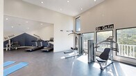 This fitness center in Memphis, TN offers a generously sized, single-floor exercise space designed to support a well-rounded workout routine. The facility features soaring vaulted ceilings that lend an open, airy feel throughout, complemented by recessed lighting and large windows on multiple walls that flood the interior with natural light and provide views of the surrounding greenery.

The exercise room is divided into functional zones. A dedicated cardio area hosts multiple treadmills positioned along a boldly accented navy blue accent wall, complete with motivational phrases such as "No Excuses" and "No Pain." A separate strength-training zone includes a multi-function cable machine, a weight bench, a leg press machine, and additional resistance equipment. Cycling enthusiasts will find stationary bikes available as well. A stretch and yoga area provides blue exercise mats laid out on the durable dark rubberized flooring, offering ample room for floor exercises and cool-downs.

The interior design balances clean white walls with striking navy blue geometric accents, creating an energizing yet modern atmosphere. The dark, low-maintenance flooring throughout provides a practical and polished finish. With wide open floor space, varied equipment options, and a bright, motivating environment, this fitness center is well-suited for a diverse range of workout preferences and fitness goals.