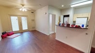 This single-floor residence offers a straightforward, functional layout centered around an open living area that connects seamlessly to the kitchen. Rich hardwood-style flooring extends throughout the main living space, adding warmth and visual continuity. A ceiling fan with a light fixture is positioned overhead, providing both airflow and ambient lighting to the room.

The front entryway features a door with a sidelight panel fitted with blinds, allowing natural light to filter in while maintaining privacy. A large HVAC return vent is mounted on the wall near the entry, and a visible thermostat or utility panel sits nearby, reflecting practical, well-integrated systems throughout the home.

The kitchen is defined by a peninsula counter with a dark granite or stone-style surface, offering additional workspace and a natural dividing point between the kitchen and living area. Light-toned cabinetry is partially visible, and what appears to be a washer or dryer unit can be seen through louvered closet doors, indicating an in-unit laundry area. A hallway leads toward additional rooms, suggesting further living or sleeping spaces beyond the main open area. The overall ambiance is clean, neutral, and move-in ready.